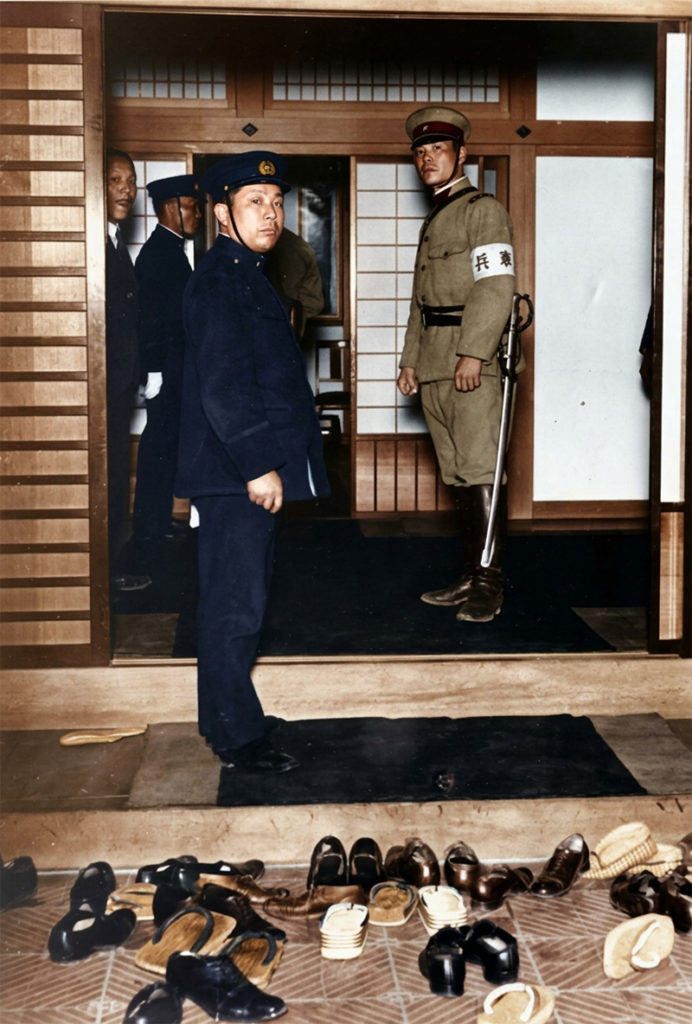 1932年、五・一五事件直後の首相官邸玄関を写したカラー化写真。警察官と「憲兵」の腕章をつけた軍人が立ち、手前には多くの靴が脱ぎ捨てられている様子。