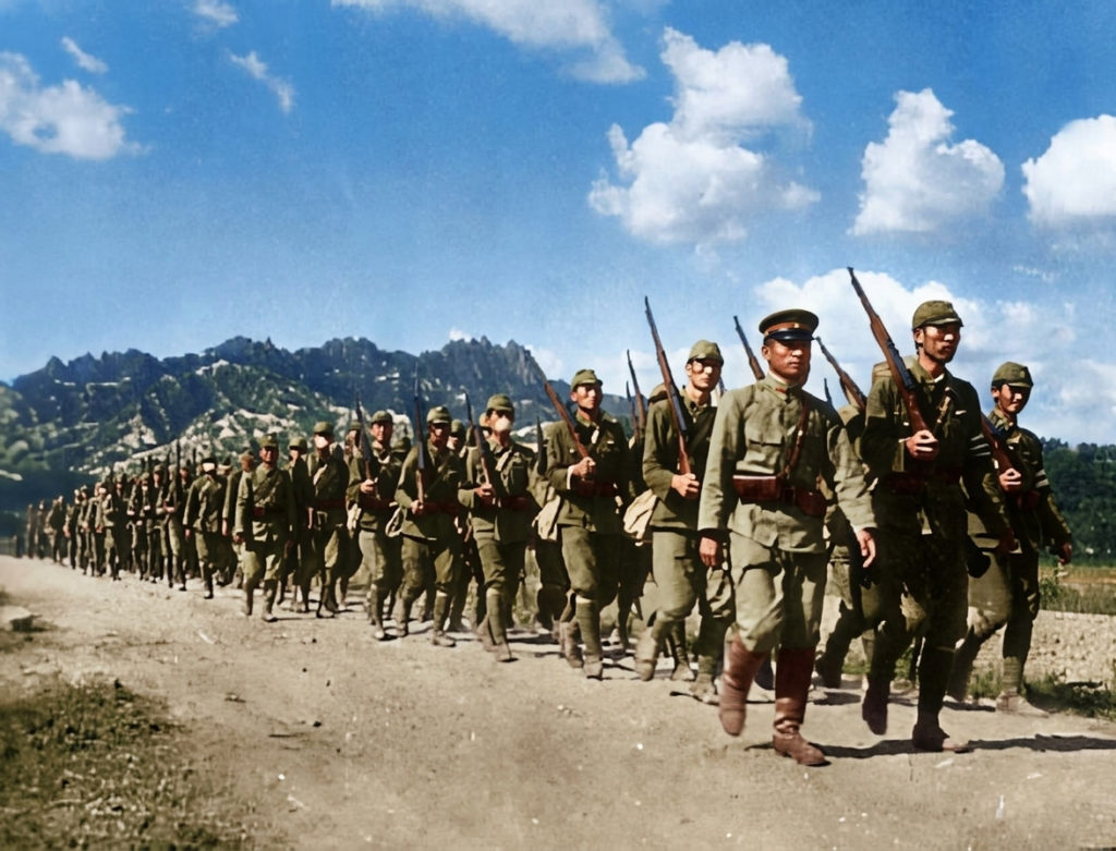 1930年代後半から1940年代にかけて、日本軍の軍服を着て銃を肩に担ぎ、山道を隊列を組んで行軍する朝鮮人志願兵たちの白黒写真（カラー化済み）。
