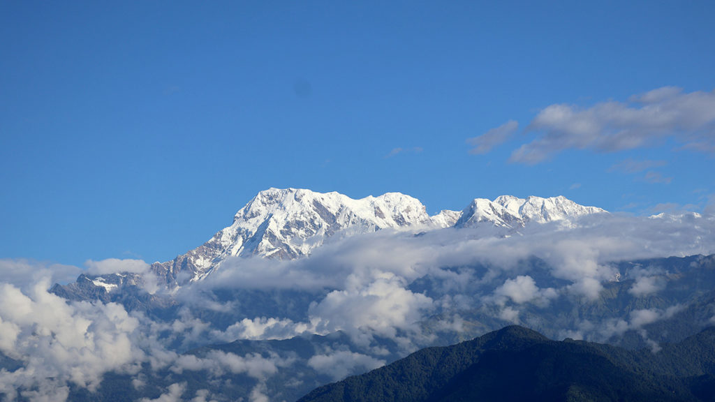 青空の下、雲海から突き出た、雪に覆われた険しいヒマラヤ山脈の高峰。手前には深い緑の山々の尾根が見える。