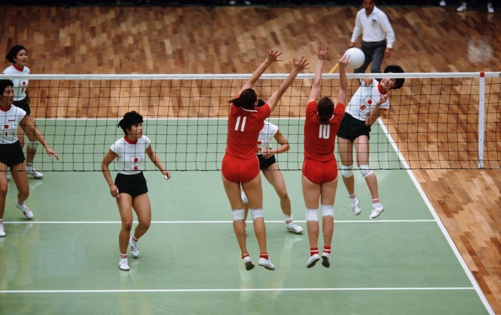 1964年東京オリンピック女子バレーボール決勝戦。ネット際でスパイクを打つ白のユニフォームの日本選手と、それをブロックしようとジャンプする赤のユニフォームのソ連選手のカラー化写真。
