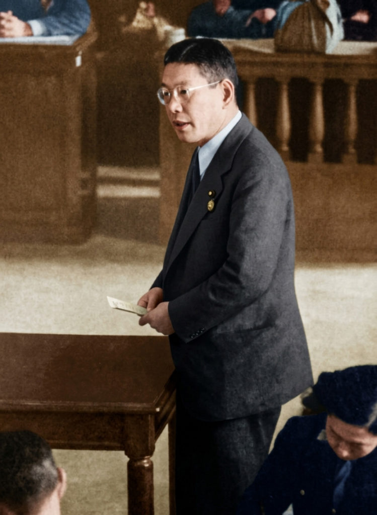 国会の演説台に立ち、眼鏡をかけて答弁する池田勇人首相のカラー化写真。国会で、手にメモを持って話している様子。