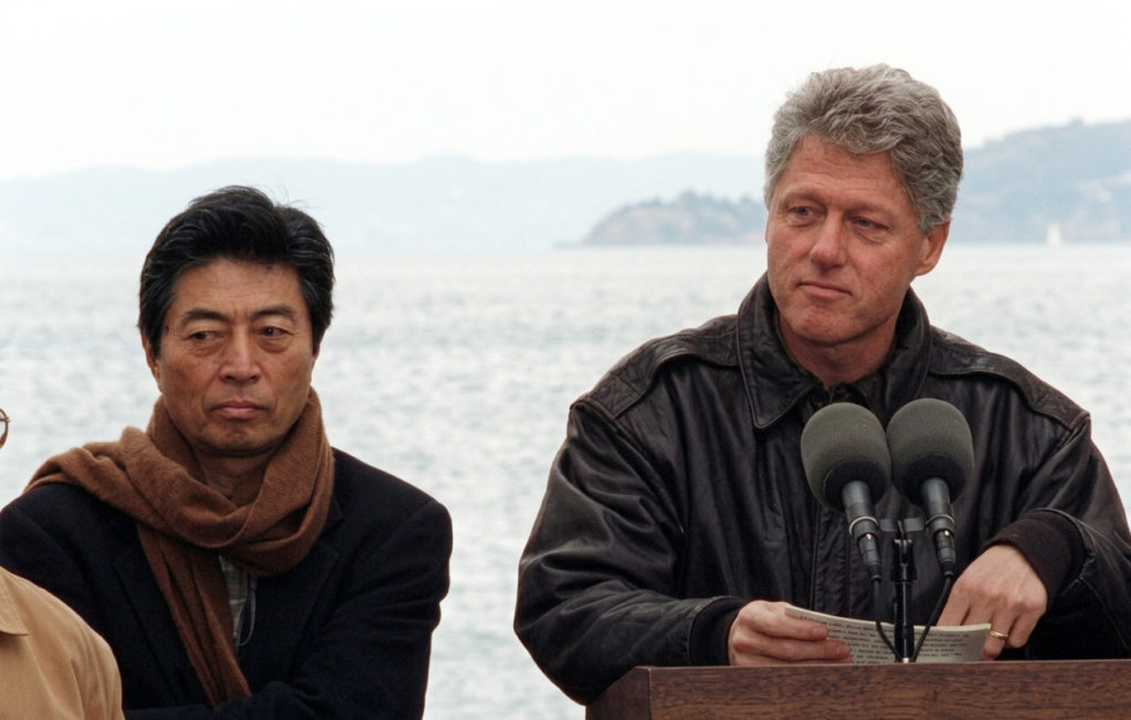 1993年のAPEC首脳会議にて、演説台に立つビル・クリントン米大統領と、その隣でマフラーを巻き穏やかな表情を浮かべる細川護煕首相。背景にはシアトルの海が広がっている。