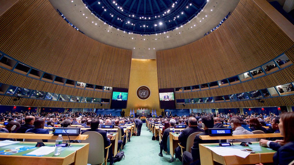 国連総会会議場の内部。ドーム型の天井と木目の壁に囲まれた広い会場に、多くの加盟国代表が着席しており、前方の壇上には大きな国連のエンブレムが掲げられている。